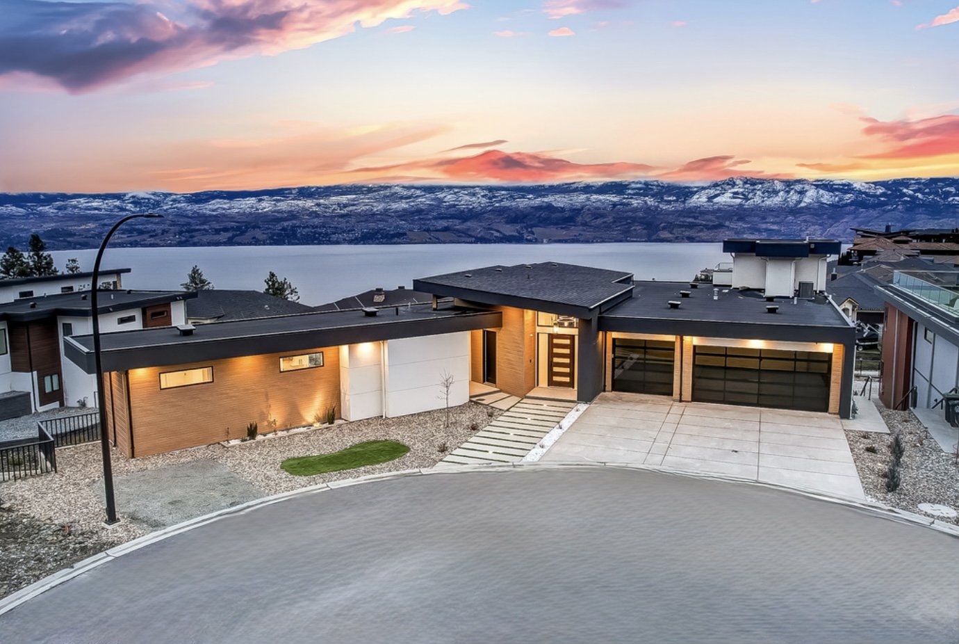 Two-storey custom home at dusk with dark cladding, a stone base, warm wood soffits, and large windows lit from inside